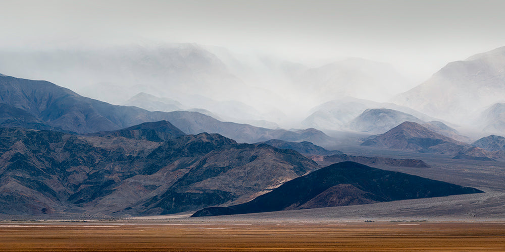 A misty mountain landscape with layered dark rocky peaks fading into a hazy gray sky, above a flat tan plain in the foreground.